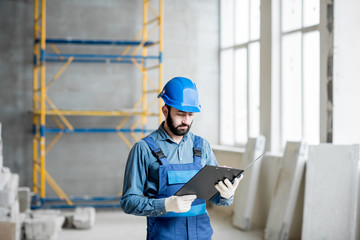 Fototapeta premium Builder or foreman in working uniform expertising the structure standing with folder at the construction site indoors