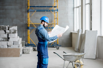 Builder or foreman in working uniform expertising the structure standing with blueprint at the construction site indoors