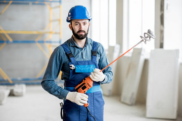 Portrait of a builder in working uniform standing with drill at the construction site indoors