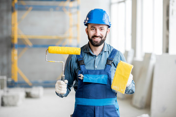 Portrait of a builder in working uniform holding yellow paint roll and grinding tool indoors