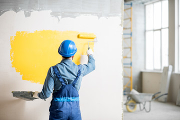 Workman in uniform painting wall with yellow paint at the construction site indoors - obrazy, fototapety, plakaty