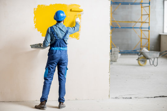 Workman in uniform painting wall with yellow paint at the construction site indoors