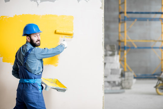 Portrait of a handsome painter in working uniform painting wall with yellow paint