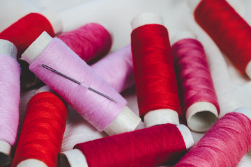 Spools of pink and red threads and sewing needle on white cotton cloth