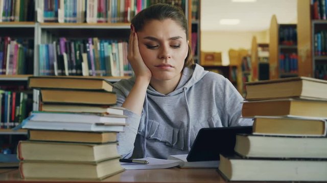 Beautiful caucasian teenage woman is sitting at table in university library holding digital tablet. She is tired and exhausted, shaking her head and writing in copybook
