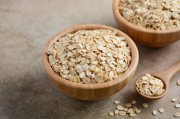 Oat flakes in a wooden bowl and wooden spoon on light brown background, selective focus.