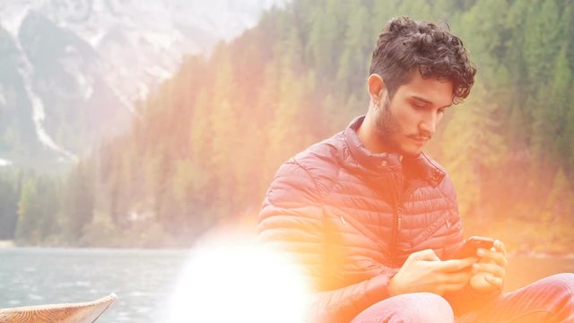 Young handsome typing text message on boat on a lake in forest, in the mountains. Braies lake or Pragser Wildsee in Trentino region, Italy