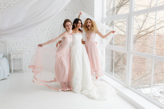 Happy bridesmaid hugging the bride covered by veil in the bedroom in the morning, having fun.