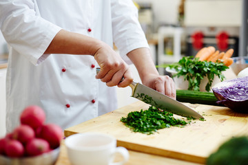Chef cutting greenery with sharp professional knife on wooden board while cooking in the kitchen