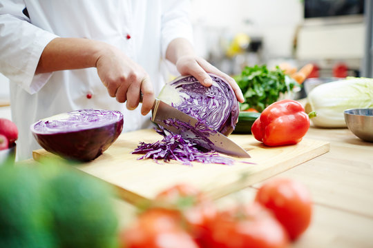 Chef in uniform cutting fresh purple cabbage on wooden board while preparing vegetable stew