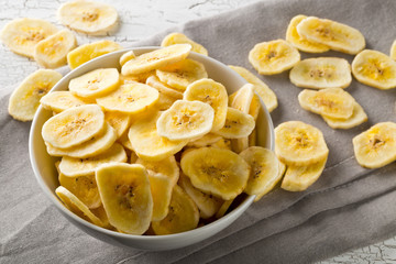 Heap of dried banana chips snack in white bowl on rustic table