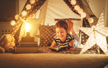 Obraz na płótnie Canvas happy child girl laughing and reading book in dark in tent at home