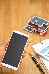 hand holding a smartphone in front of house model,calculator,pen,and coins on wooden table