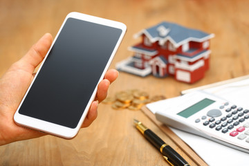 hand holding a smartphone in front of house model,calculator,pen,and coins on wooden table