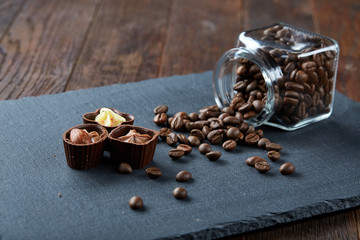 Side view of overturned glass jar with coffee beans and chocolate candies on wooden background, selective focus