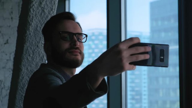 young successful businessman doing selfie on the background of a window overlooking the downtown. High business building. Office in loft style.