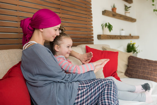 Young adult female cancer patient spending time with her daughter at home, relaxing on the couch. Cancer and family support concept.