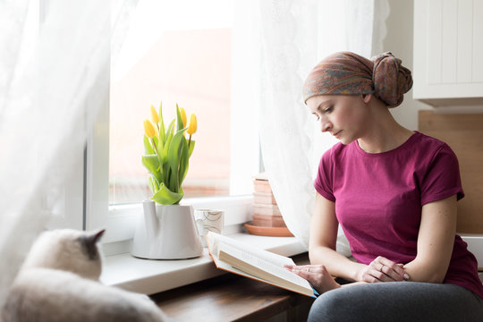 Young Adult Female Cancer Patient Wearing Headscarf Sitting In The Kitchen With Her Pet Cat, Relaxing By Reading A Book.
