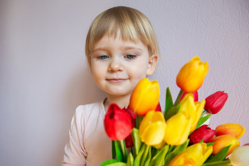 Fototapeta na wymiar Happy little girl with blond hair holding a bouquet of colorful tulips in hands in a sunny room in the house. Holy concept.