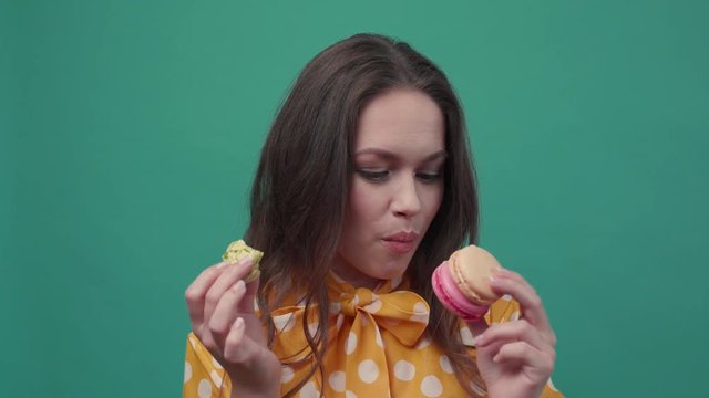 Pretty young woman in orange blouse happily eats the macaroons, choosing between colorful ones and green cookie, than looks straight to camera and raises her eyebrows. True emotions, having fun. Slow