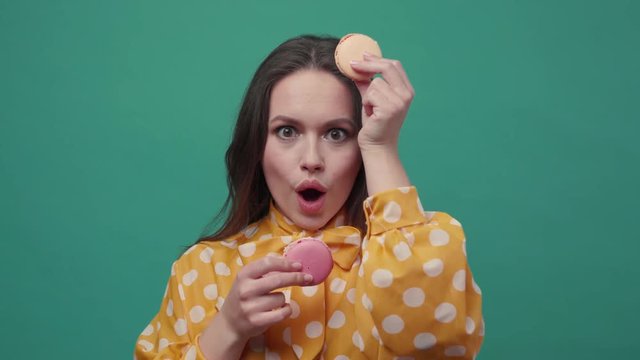 Hilarious young woman with comical face funnily gesticulates with two colorful macarons on a dark green background. Happy, cheerful mood. Delicious, sweet tooth. Slow motion, close up view, female
