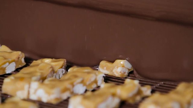 Bars of nougat with nuts are poured with chocolate. Manufacture of chocolate bars with nougat and peanuts. Chocolate bars move along conveyor belt at confectionery factory for the production of sweets