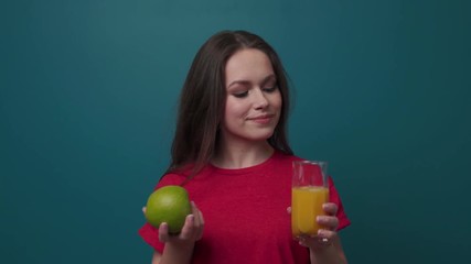 Attractive brunette woman in casual look chooses between green apple and a glass of orange juice, charmingly smiles and drinks the juice. Diet, healthy lifestyle, fit figure. Close up, slow motion - Powered by Adobe