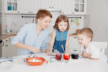 Mother, daughter and son paint eggs. White kitchen. A happy family is preparing for Easter.