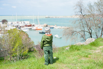 little boy in jacket and hat stands on green hill on background of sea bay with ships in early spring