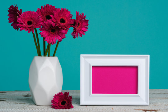 Happy Mother's Day, Women's Day, Valentine's Day or Birthday Pastel Candy Blue Coloured Background. Dark pink gerberas in a vase on a table with empty picture frame greeting card.