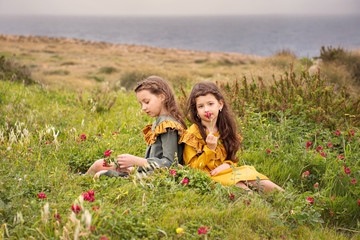 two sister girls in old-fashioned vintage dresses sniffing flowers sitting on a plateau near the stormy sea