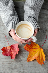 A cup of coffee in hands and autumn leaves on a wooden table
