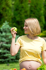 A pregnant woman in a yellow T-shirt sitting on grass and holding an apple in hand and at the bottom stands a tray of fresh fruits and vegetables . Rest at nature. Fresh air and healthy food. The