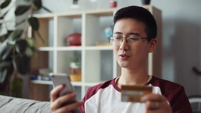 Serious chinese young man with glasses sit on sofa use phone and credit card at home shopping internet asian online boy korean mobile shop smart text cellphone businessman credit card buy portrait