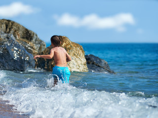 Cute Caucasian boy is running in the water along the sea shore. Back view.