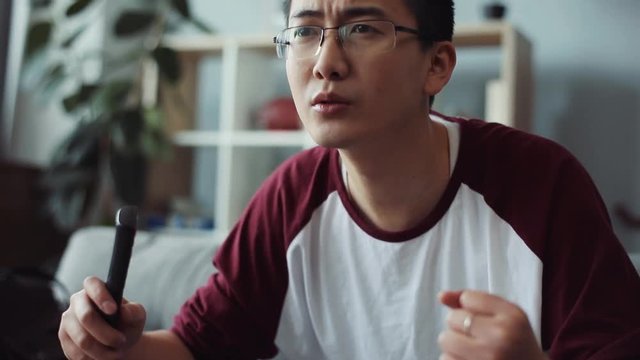 Close up face smiling attractive young chinese man with glasses sits on the couch and turns on the TV at home looking house technology male business bored leisure video sofa portrait slow motion