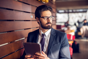 Portrait of young thoughtful handsome bearded businessman in suit listening music from a tablet at a modern cafe.