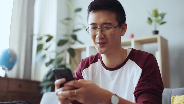 Happy chinese young man with glasses sit on sofa use phone at home man business house internet technology asian cell cellphone happy boy male online smartphone gadget slow motion portrait close up