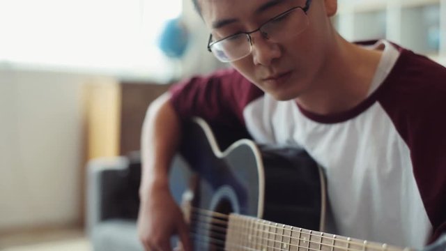 Concentrated handsome asian young man playing guitar sit on sofa at home acoustic asian casual art melody musician song attractive audience band boy coast artist sound portrait close up slow motion