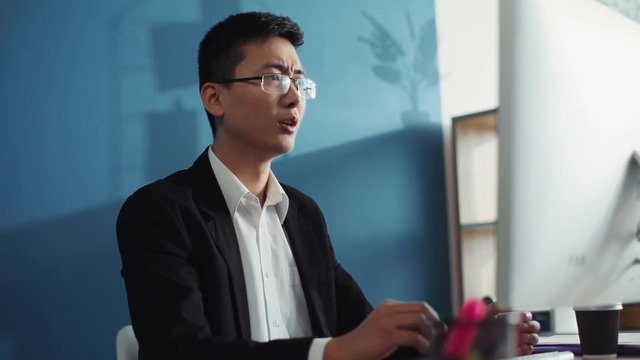 Nervous young chinese man with glasses uses computer to look at the screen in office business internet face technology businessman laptop male sitting job attractive interior handsome slow motion