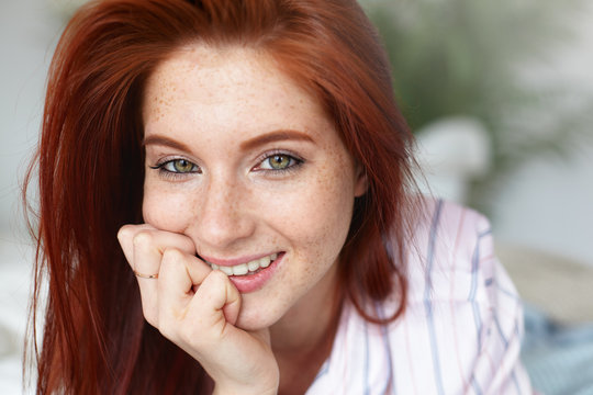 Picture of charming young female with loose red hair and freckles lying on bed at home, keeping hand under her chin and looking at camera with happy positive smile, posing against blurred background