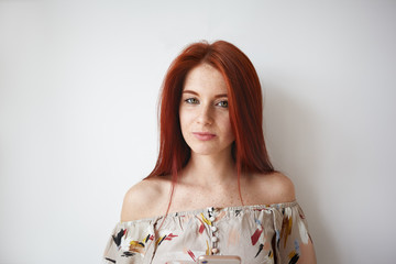 Cute young female with loose red hair using cell phone posing isolated against white blank wall with copy space for your information, dressed in romantic summer top with off the shoulder design