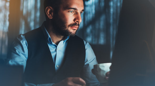 Bearded young businessman working on modern office. Consultant man thinking looking in monitor computer. Manager typing on keyboard in coworking workplace, startup project concept in night with coffee