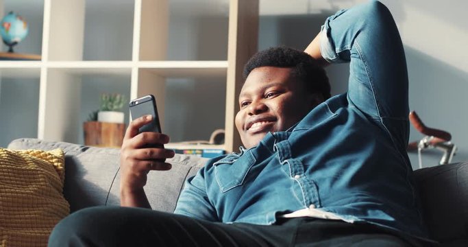Bored young african american man use phone lazy sit on sofa in home online relax work cell phone app authentic bills sunset guy close up mobile smartphone slow motion portrait handsome rude