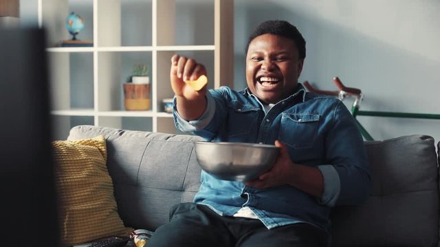 Happy rude african american man watching tv sit on couch eat chips smile laughs changing channel commercials control flipping guy handsome cute home interior technology portrait slow motion