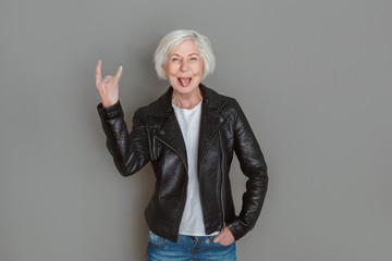 Senior woman in leather jacket studio isolated on grey wall tongue out showing rock on