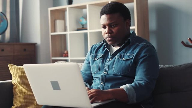 Unhappy young african american handsome man uses computer work in home sit on sofa programmer looks at monitor screen freelance successful worker businessman engineering Slow motion portrait close up