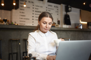 People, business, rest and modern technology concept. Picture of serious middle aged businesswoman in white formal shirt sending email using laptop while having hot drink at cozy coffee house