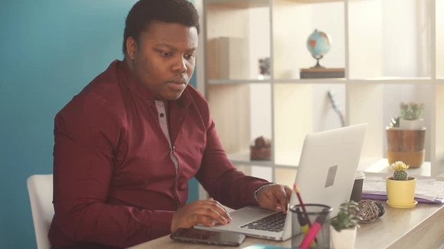 Young african american man thinking and working on laptop typing in office sunlight business person internet face businessman freelance looking portrait serious work slow motion black brainstorming