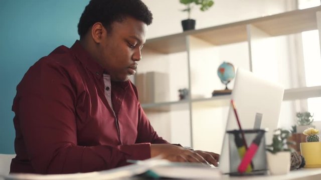 Young african american man thinking and working on laptop holds documents in office sunlight business internet face businessman freelance looking portrait serious work slow motion black brainstorming
